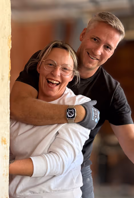 Lächendes Paar, der Mann umarmt die Frau von hinten, beide tragen lässige Kleidung.